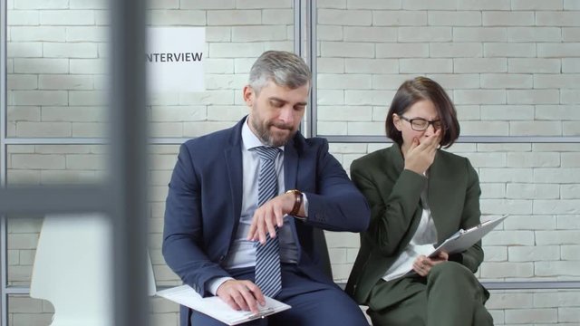 Tracking shot of annoyed bearded man in suit and yawning young woman in glasses sitting in chairs in office reception area and complaining while waiting for job interview