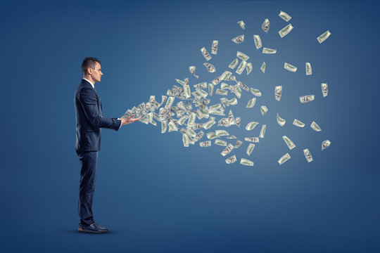 A single businessman stands in a side view with many USD dollar banknotes flying out from his hands.