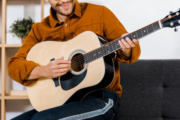 cropped view of cheerful man in glasses playing acoustic guitar at home