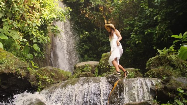 Attractive Girl in White Dress and Straw Hat Walking Barefoot near Small Waterfall in Tropical Rainforest Jungle. Carefree Lifestyle Travel 4K Slowmotion Footage. Bali, Indonesia.