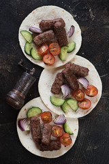 Tortilla flatbread with bbq cevapcici or skinless beef sausages and vegetables, above view on a dark brown stone surface