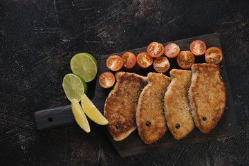 Roasted fish cutlets with cherry tomatoes, lemon and lime. Flatlay on a dark brown stone background, horizontal shot