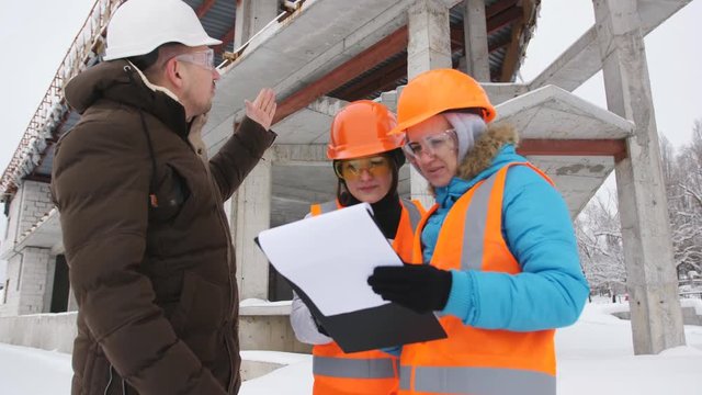 The foreman and two women inspector in the winter, inspect the construction site located behind them in the background.