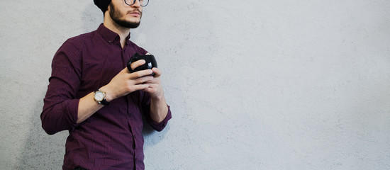 Young bearded hipster guy with black cup of coffee in hands. With hat on the head and wearing glasses. On background of white.