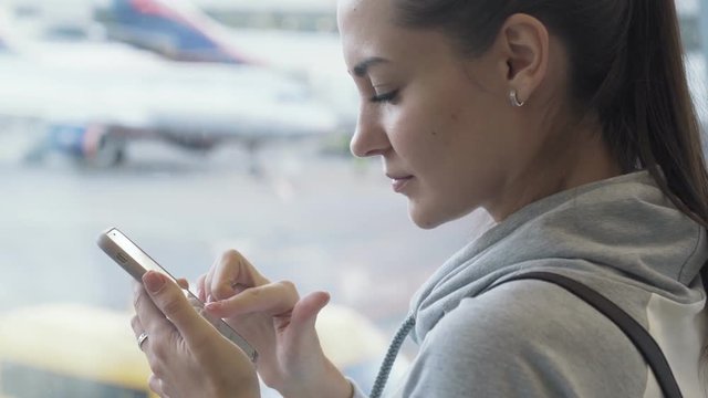 Side view attractive girl using smartphone and texting message in airport background of plane in window, close-up