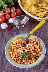 Boiled chickpeas, seasoned with vegetable oil, ground red pepper, sesame seeds and onion rings in a bowl with old spoon on a wooden background next to garlic and tomatos and corn chips, vertical orien