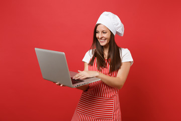 Housewife female chef cook or baker in striped apron, white t-shirt, toque chefs hat isolated on red wall background. Fun housekeeper woman looking for recipe in laptop pc. Mock up copy space concept.