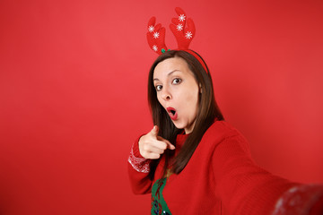 Close up selfie shot of amazed Santa girl in decorative deer horns pointing finger on camera, keeping mouth wide open isolated on red background. Happy New Year 2019 celebration holiday party concept.