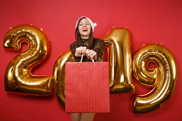 Merry Santa girl in shiny glitter dress, Christmas hat hold shopping bag isolated on bright red wall background, golden numbers air balloons studio portrait. Happy New Year 2019 holiday party concept.