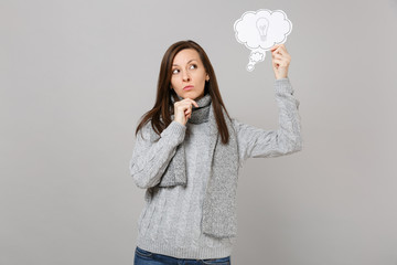 Pensive young woman in gray sweater, scarf put hand prop up on chin, hold say cloud with lightbulb isolated on grey background. Healthy fashion lifestyle, people sincere emotions, cold season concept.
