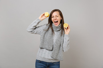 Cheerful young woman in gray sweater, scarf blinking, holding lemons isolated on grey background in studio. Healthy fashion lifestyle, people sincere emotions, cold season concept. Mock up copy space.