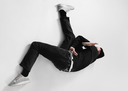 Freestyle Dancer Dressed In Black Jeans, Sweatshirt, Hat And Gray Sneakers Is Dancing Lying On The Floor In The Studio On The White Background