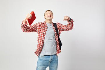 Fototapeta premium Joyful blond guy with black backpack on his shoulder dressed in a white t-shirt, red checkered shirt and jeans holds book in his hand on the white background in the studio