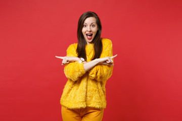 Excited young girl in yellow fur sweater with opened mouth holding hands crossed pointing index fingers aside isolated on red background. People sincere emotions lifestyle concept. Mock up copy space.