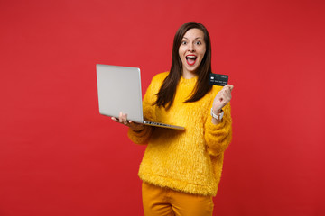 Excited young woman in yellow fur sweater keeping mouth wide open, hold credit bank card, laptop pc computer isolated on red background. People sincere emotions, lifestyle concept. Mock up copy space.