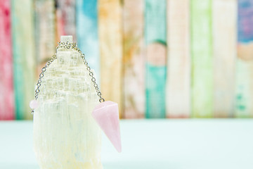Using crystal Selenite tower for loading rose quartz pendulum, light blue table and pastel color wooden board background. Divination and fortune telling concept.