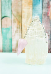 Using crystal Selenite tower for loading rose quartz pendulum, light blue table and pastel color wooden board background. Divination and fortune telling concept.
