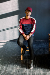 Portrait of happy young black African guy in red baseball cap sitting in a studio on a wooden chair on a dark background .