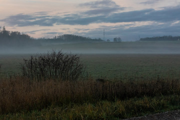 Obraz premium sunrise in the countryside, fog on the field with small hills, small groves in the distance; The sky is yellow with blue clouds