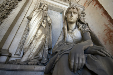 Genoa / Italy - June 20 / 2016 : Statue of two women, one is standing in front of a door orher is sitting thoughtfully at monumental cemetery of Staglieno