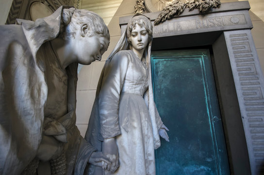 Genoa / Italy - June 20 / 2016 : Statue of two women, one holding other woman's hand in front of a door at monumental cemetery of Staglieno
