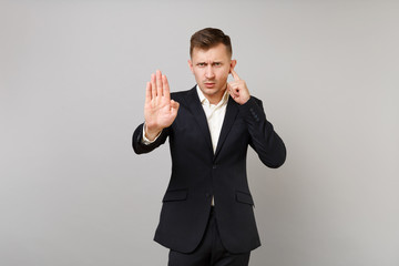 Concerned young business man in classic black suit covering ear with finger, showing stop gesture with palm isolated on grey background. Achievement career wealth business concept. Mock up copy space.