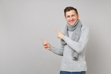 Attractive young man in gray sweater, scarf pointing index fingers aside isolated on grey wall background. Healthy fashion, lifestyle people sincere emotions, cold season concept. Mock up copy space.