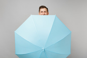 Smiling young man in gray sweater, scarf hiding behind blue umbrella isolated on grey background in studio. Healthy fashion lifestyle people sincere emotions, cold season concept. Mock up copy space. © ViDi Studio