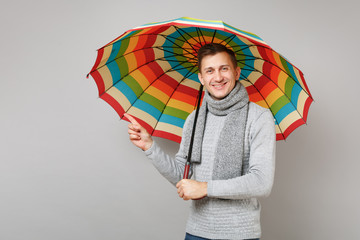 Smiling young man in gray sweater, scarf holding colorful umbrella, pointing index finger aside...