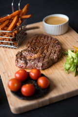 A platter of sirloin steak, salad, fries, sauce, tomato and mushrooms, served on a rustic wooden cutting board. Dark food photography with a mechanics workshop industrial setting.