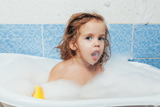 Fun cheerful happy toddler baby taking a bath playing with foam bubbles. Little child in a bathtub. Smiling kid in bathroom on blue background. Hygiene and health care.