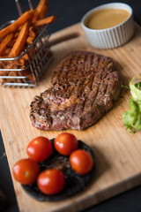 A platter of sirloin steak, salad, fries, sauce, tomato and mushrooms, served on a rustic wooden cutting board. Dark food photography with a mechanics workshop industrial setting.
