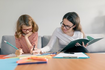 Young female teacher giving private lesson to child, little girl sitting at her desk writing in...