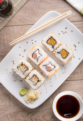 Sushi on a white square plate with soy sauce and natural wooden chopsticks on a light wooden table.