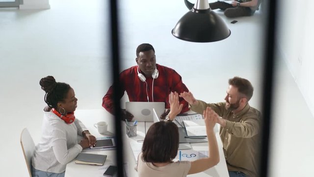 High angle tracking shot of happy multi-ethnic team of businesspeople sitting at table in meeting room and high-fiving each other after successfully finishing project