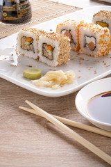 Sushi on a white square plate with soy sauce and natural wooden chopsticks on a light wooden table.