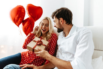 Couple. Love. Valentine's day. Emotions. Man is giving a gift box to his woman, both smiling; at home - obrazy, fototapety, plakaty