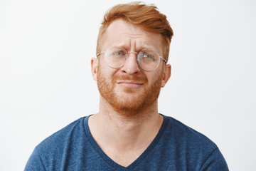 Handsome gloomy redhead male with beard frowning and pursing lips looking right with regret and disappointment, standing over gray background in casual clothes and glasses