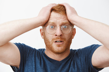 Headshot of worried attractive redhead businessman in glasses and blue t-shirt holding hands on hair and frowning, standing in stupor at shocking scene, feeling anxious and desperate