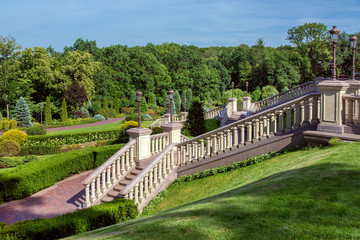 A staircase with stone railing balustrades and retro lanterns on pedestals against the backdrop of a landscape with a boxwood hedge and a forest with trees.