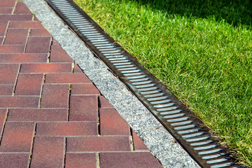 Drainage system with iron mesh along the pedestrian sidewalk with red tiles and green lawn,...