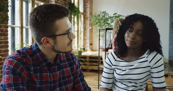 Young Caucasian man in glasses and African American woman with curly hairtalking and sharing their ideas over the sturup business project. Close up. Indoors.