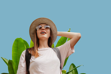 Astonished young female in stylish sunglasses and hat looking away while standing near exotic plant...