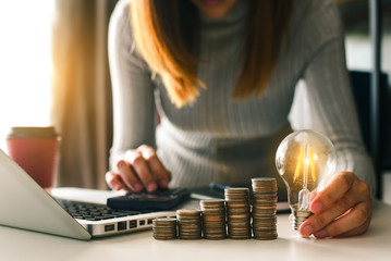 business hand holding lightbulb with using laptop computer and money stack in office. idea saving...