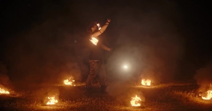 Cool male fire show stuntsman performing, flailing his burning sticks, standing in circle of burning bowls- slow motion 4k