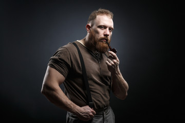 Fototapeta na wymiar Lumberjack brutal red beard muscled man in brown shirt with smoking tube standing on dark background
