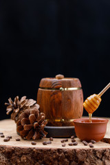 A barrel with honey and a clay cup with a honey stick on a large wooden saw cut surrounded by pine cones on a dark background. Copy space. Space under your text.