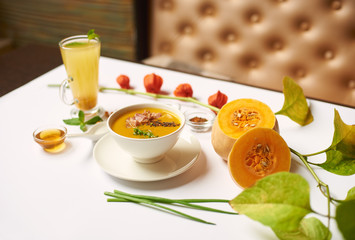 Creative still life served on the table for dinner. Delicate cream soup and tea in high cup prepared in oriental restaurant. Green leaves and halves of pumpkin lying near meal on white tablecloth.