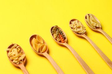 Spoons with assortment of uncooked pasta on color background