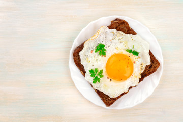A sunny side up fried egg on a bread toast, shot from above on a pale wooden background with a place for text